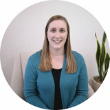 Woman seated in a calm, neutral space, smiling at the camera with a minimalist background and indoor plant, representing a trusted healthcare professional.